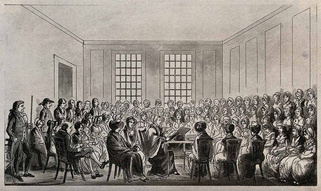 Elizabeth Fry sentada en una mesa en una gran sala rodeada de hombres y mujeres presos que la escuchan
